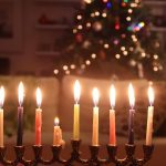 A photograph of a lit menorah with a Christmas tree in the background.