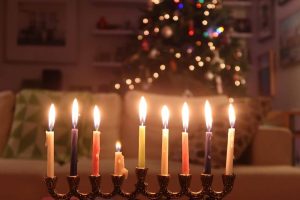 A photograph of a lit menorah with a Christmas tree in the background.